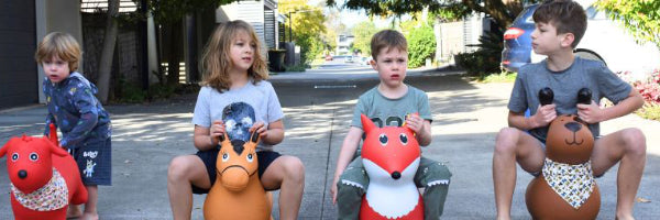 Four children riding inflatable animal bouncers outdoors in a suburban setting.