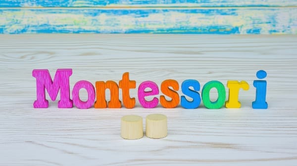 Colorful wooden letters spelling "Montessori" with two small wooden cylinders in front, on a light wooden background.