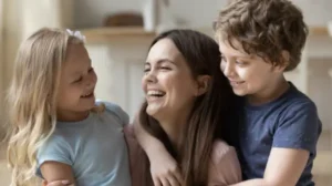 Smiling woman with two happy children hugging her, one girl and one boy, indoors.