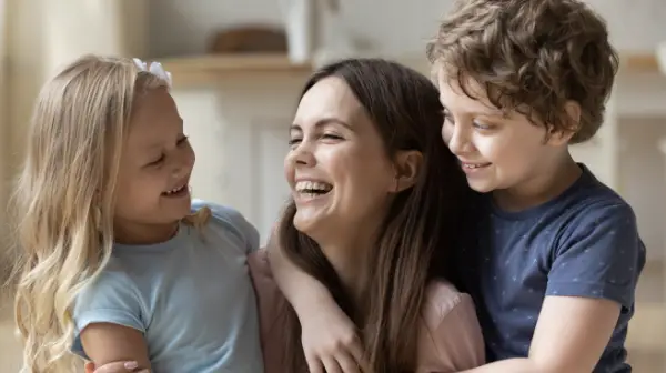 Smiling woman with two happy children hugging her, one girl and one boy, indoors.